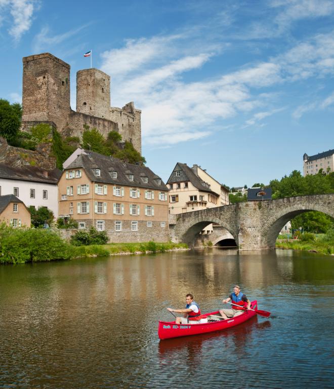 Kanu mit zwei Personen auf einem Fluss. Im Hintergrund ist eine steinerne Brücke und ein altes Dorf. Auf einem Hügel dahinter ragt eine Burg hervor.